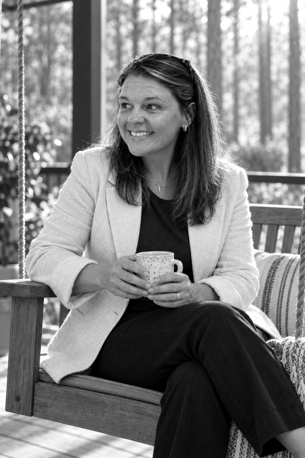 Black-and-white photo of Tara on a porch swing holding a mug, pine trees behind her